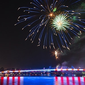 A bridge on the Arkansas river lit up in red, white, and blue with fire works going off above it during the night.